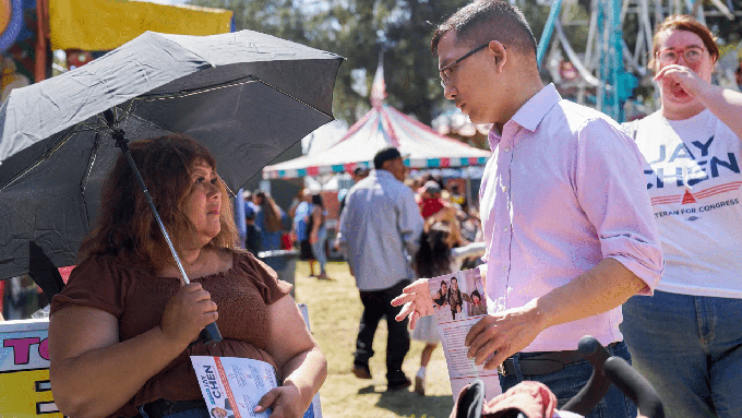 Image caption: Democratic candidate ,Jay Chen speaks with voter Ramona Mejia in Spanish while canvassing for voters at the 62nd Garden Grove Strawberry Festival on May 29, 2022.