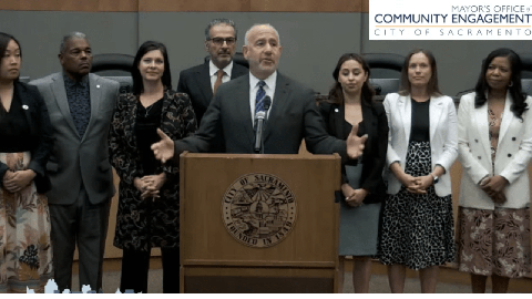 Image caption: Sacramento Mayor Darrel Steinberg stands at a podium surrounded by fellow elected officials and staff members.