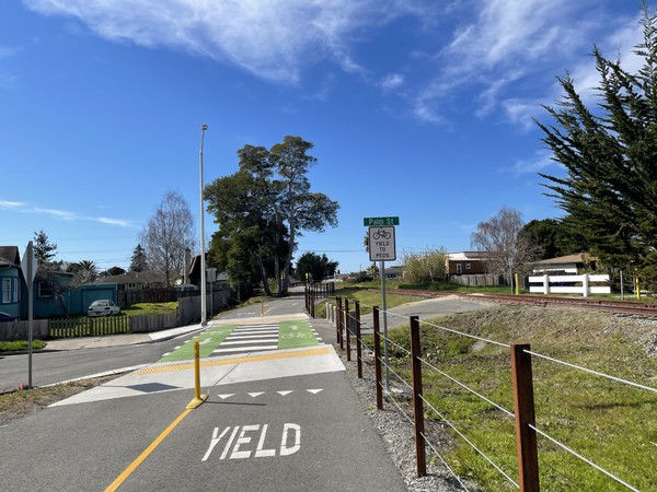 Looking east toward the intersection of the rail trail with Palm Street. 
Fun fact:  the rail here is on a raised hump, and cars used to drive down Palm Street so fast that they would hit the hump like a ramp and gain air before crashing down at the top, throwing sparks. 
There were gouges at the top of the hump from bolts on the underside of cars digging into the pavement.
In the 80's, neighbors campaigned and the city blocked off Palm Street at both sides of the tracks.