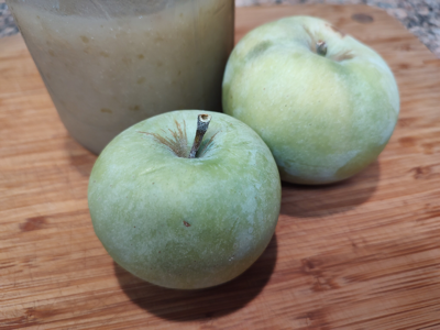 Two green apples and a container of applesauce on a wooden surface