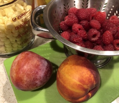 A glass measuring cup full of cereal, a metal colander with raspberries, and a pluot and a nectarine on a green cutting board