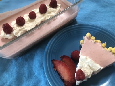 On a blue background, a glass loaf pan with pink filling and a slice of pink pie with whipped cream and fruit