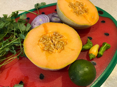 Halved melon, half a red onion, a lime, cilantro and peppers on a red cutting board