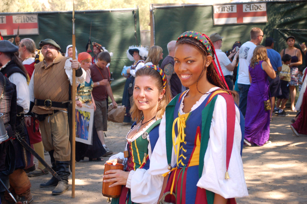 Two women in period costume pose at a crowded Renaissance fair